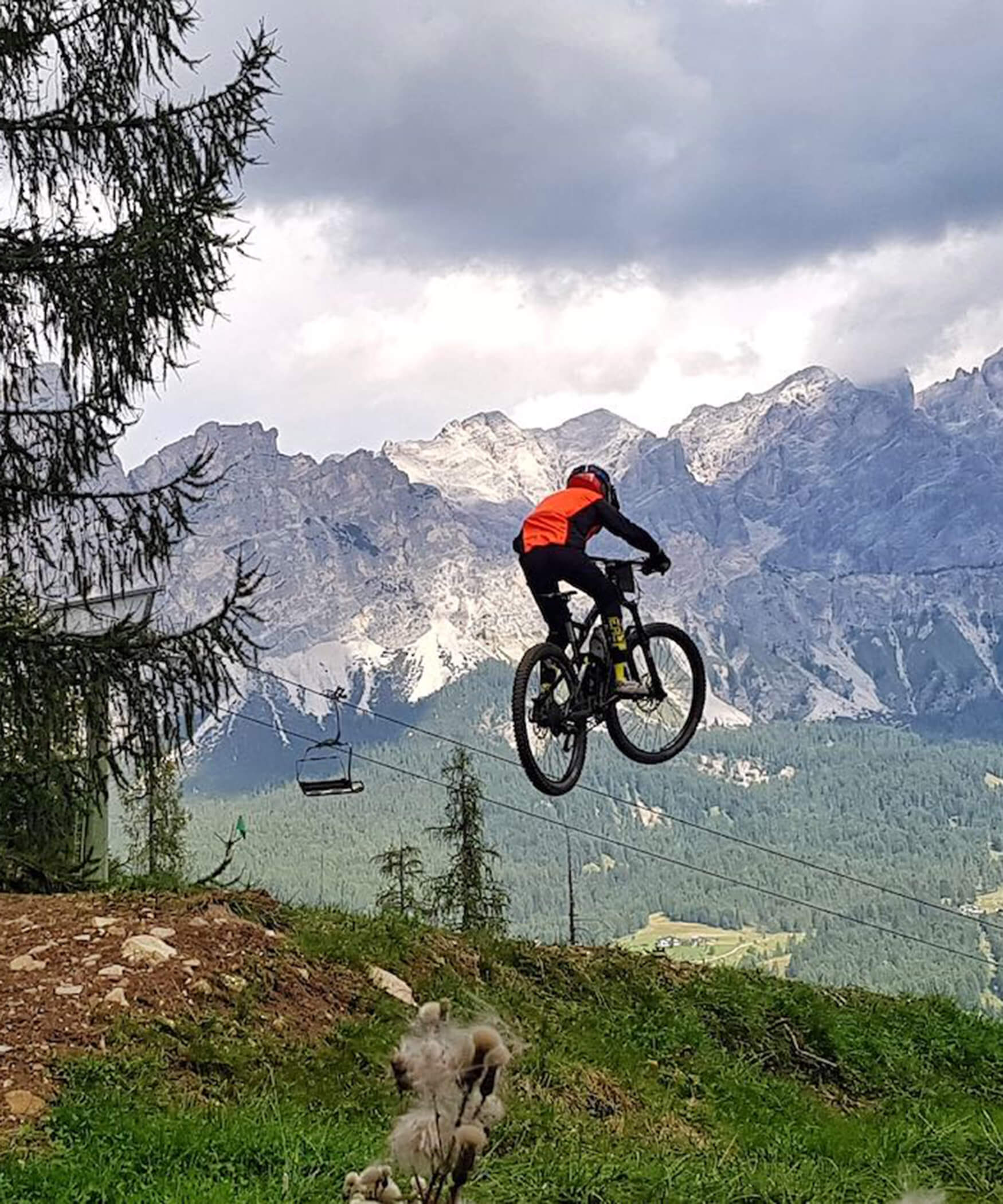 A cyclist jumps a stretch of meadow with his MTB, with a chairlift and the Dolomites in the background - Laurin's