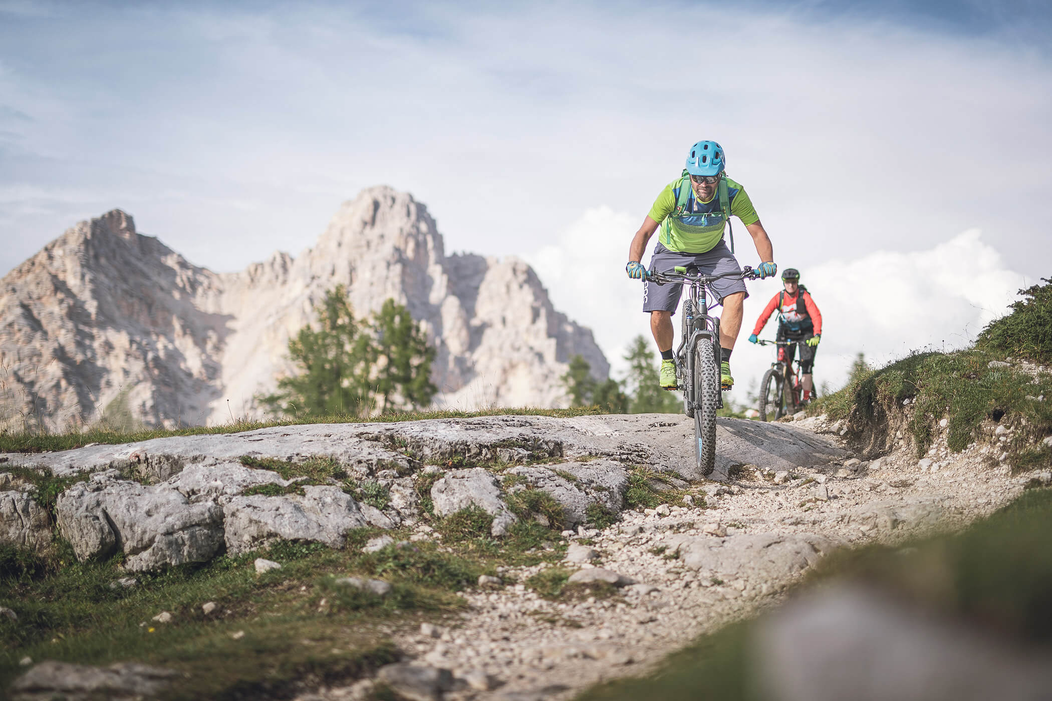 Two cyclists on a tour in the Dolomites in fine weather, with a peak and treetops behind them - Laurin's