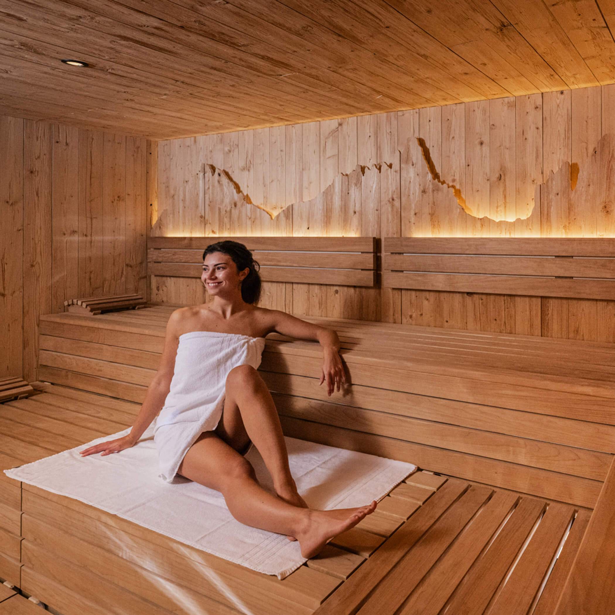 Frau sitzt in der modernen finnischen Sauna und lächelt, auf der Holztäfelung dahinter ist eine Berglandschaft eingefräst, welche indirekt beleuchtet wird - Laurin's