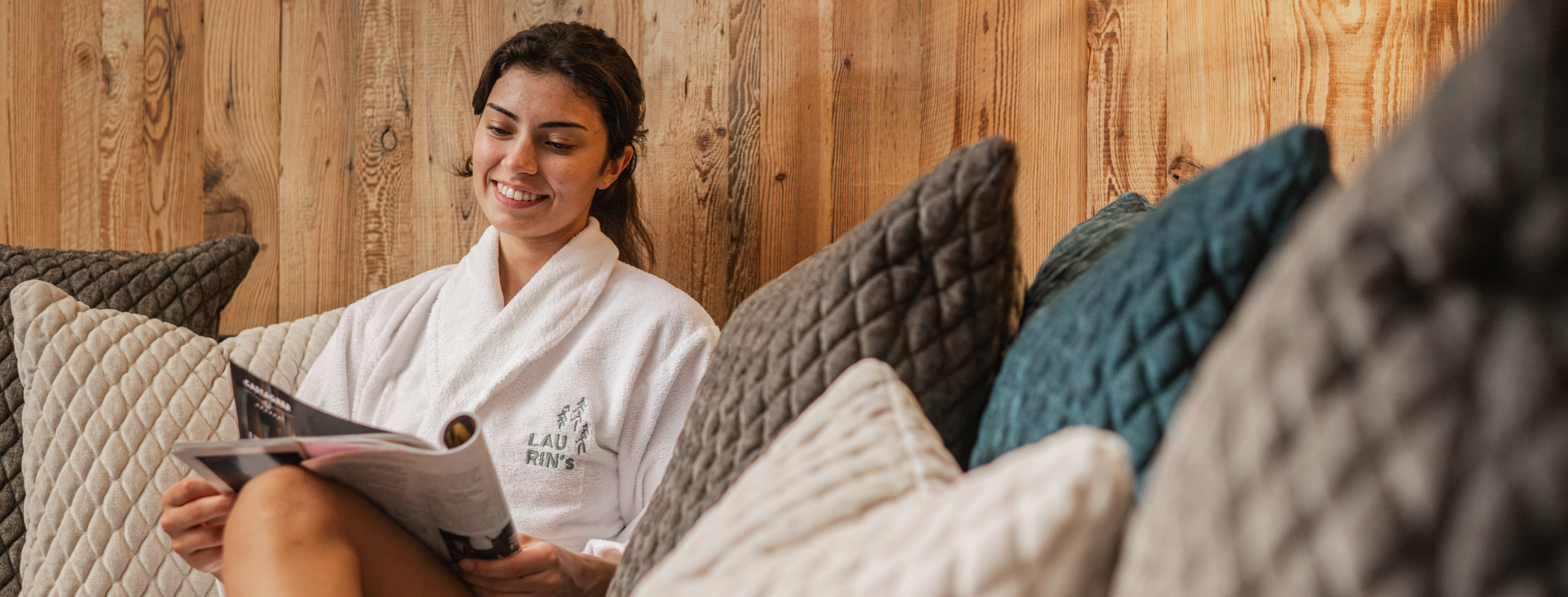 Eine Frau im Bademantel sitzt zwischen mehreren Kissen und liest ein Magazin - Laurin's